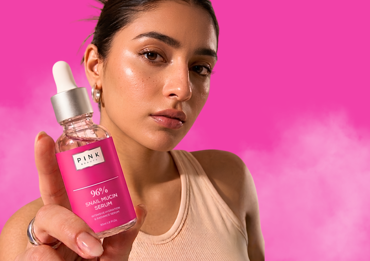 A woman holds a bottle of PINK Beauty 96% Snail Mucin Serum against a vibrant pink background. The woman is wearing a beige ribbed tank top and holding the product bottle in her hand.