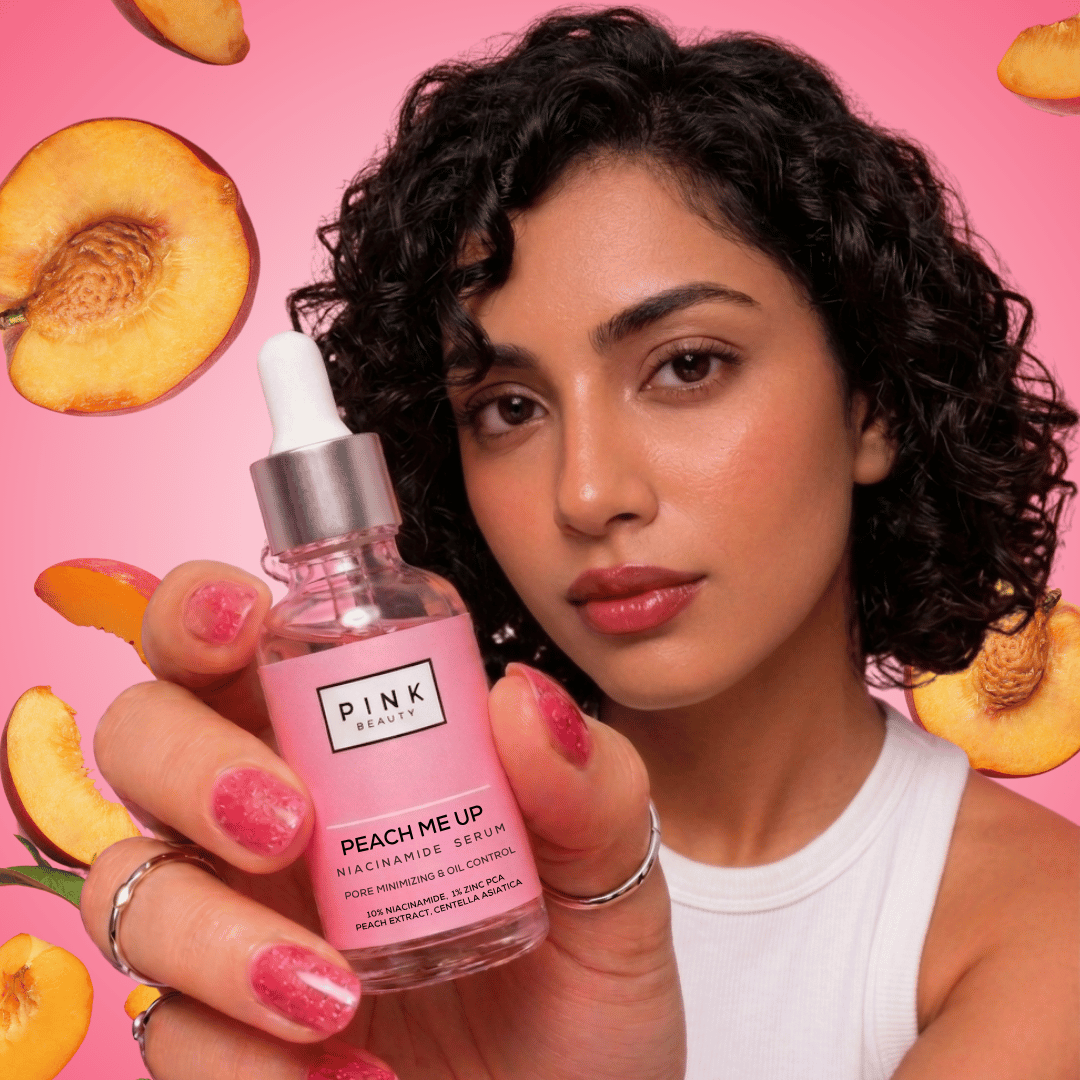 A woman holding a bottle of PINK Beauty Peach Me Up Hyaluhydrating Serum against a pink background with floating peach slices. The bottle is clear glass with pink liquid, a white dropper cap, and a silver collar.