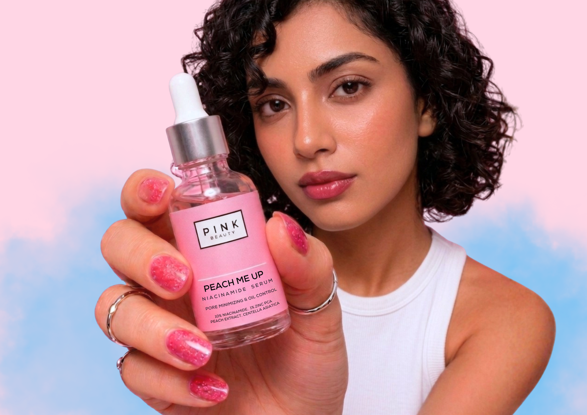 A woman holding a bottle of PINK Beauty 'Peach Me Up' serum against a pastel pink and blue background. The woman has curly dark hair, is wearing a white tank top, and has glittery pink nails.