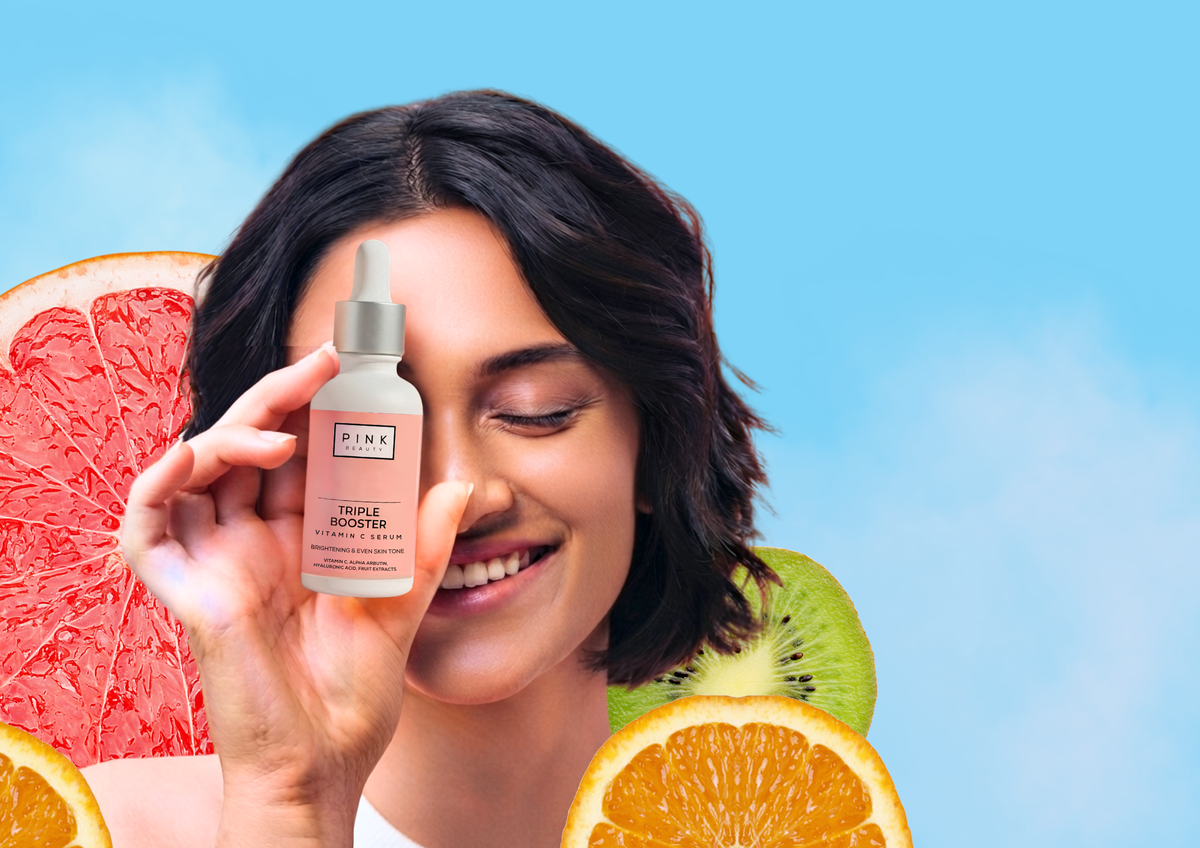 A woman holds up a bottle of PINK Beauty Triple Booster Vitamin C Serum against a bright blue background with stylized fruit slices including grapefruit, kiwi, and orange.